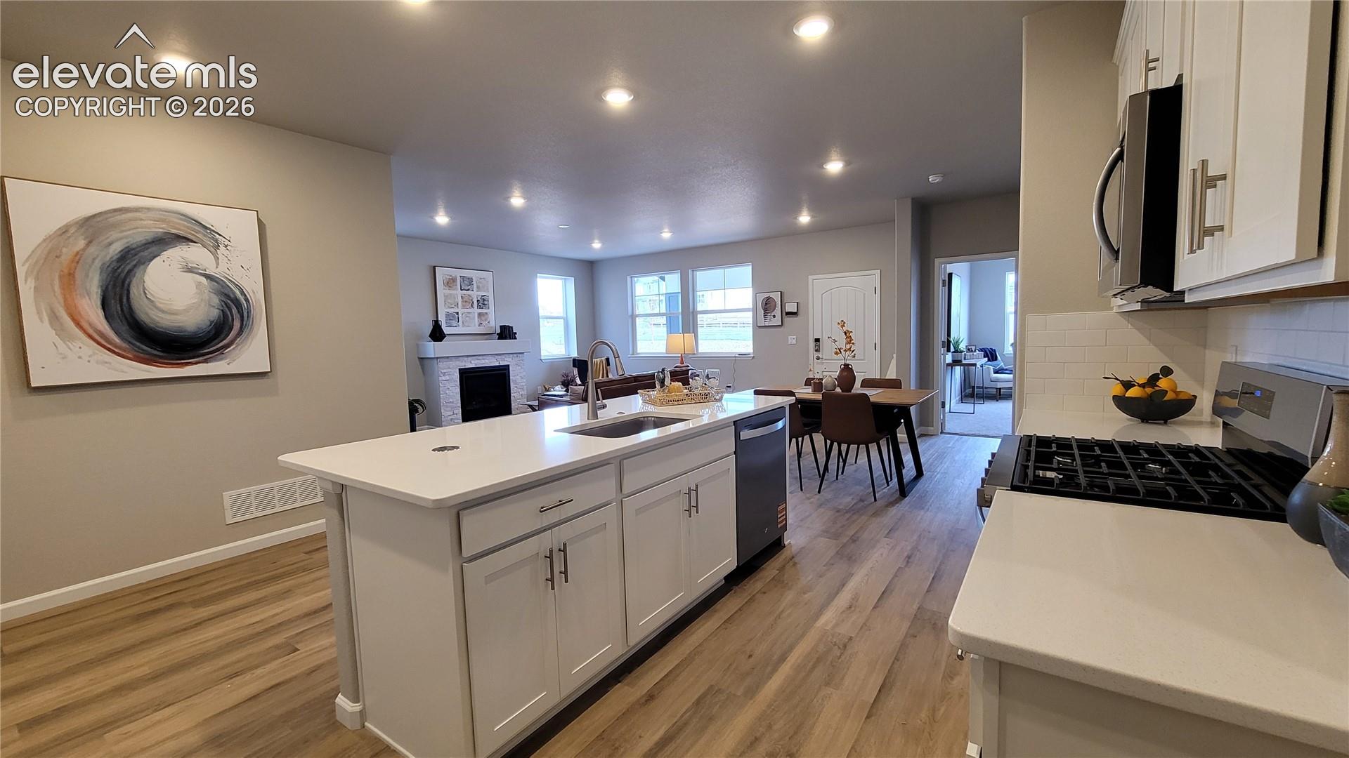 Kitchen featuring white shaker-style cabinetry, quartz countertops, a central island with a stainless steel sink, LED lighting, and LVP flooring, pantry with wood shelving, and stainless steel gas range, microwave, and dishwasher!