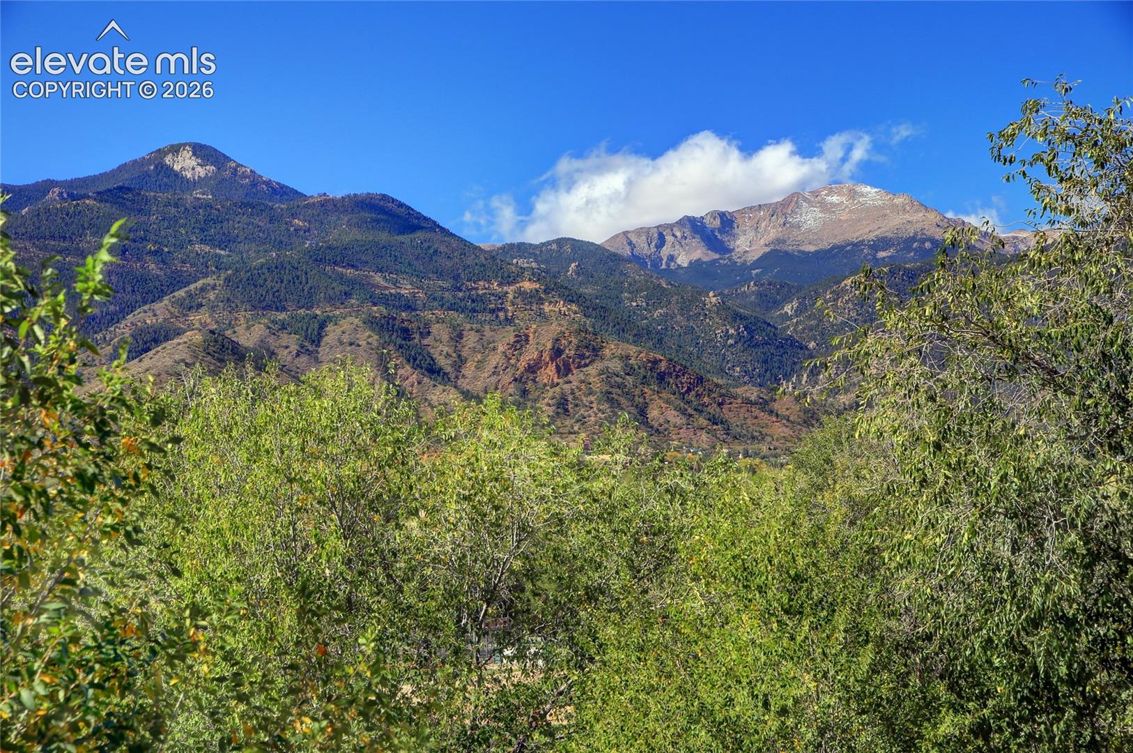 Mountainous backdrop featuring prominent rocky peaks, dense evergreen forest, and lush deciduous foliage