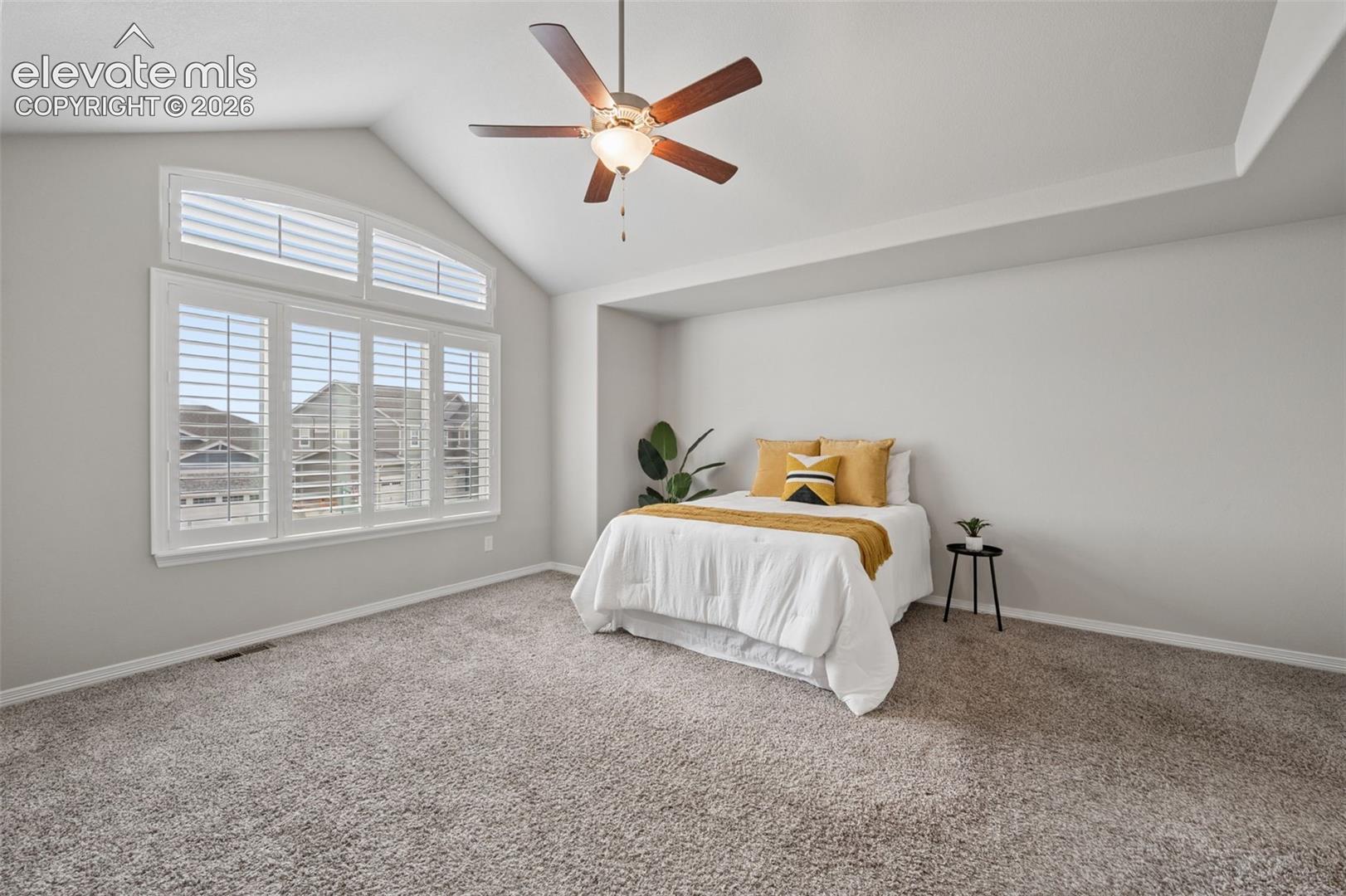 Vaulted ceiling with a ceiling fan, tray ceiling detail, large window with white plantation shutters, and light grey wall finishes