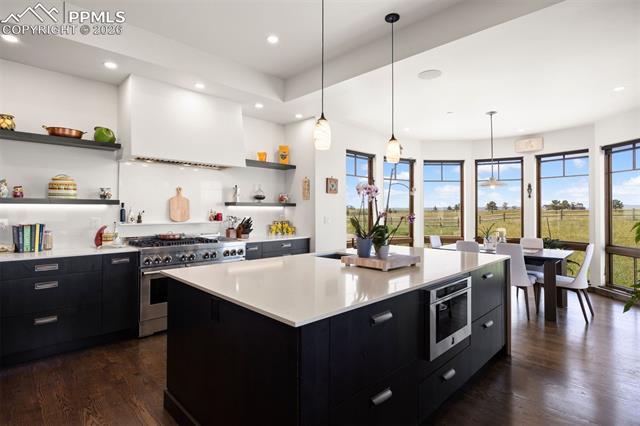 JennAir gas range with custom hood, open shelving, and eat-in area surrounded by panoramic windows.