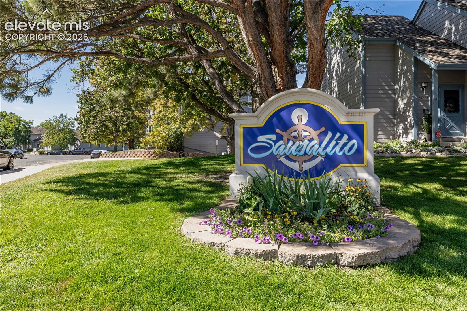 Community signage featuring a stone-based monument with integrated landscaping, manicured lawn, and mature canopy trees