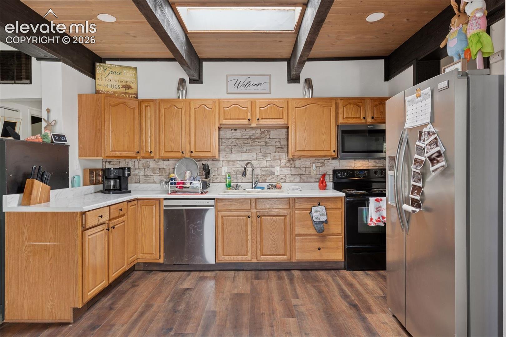 Kitchen featuring wood cabinetry, stone-like tile backsplash, stainless steel appliances, and vaulted ceiling with exposed beams and skylight
