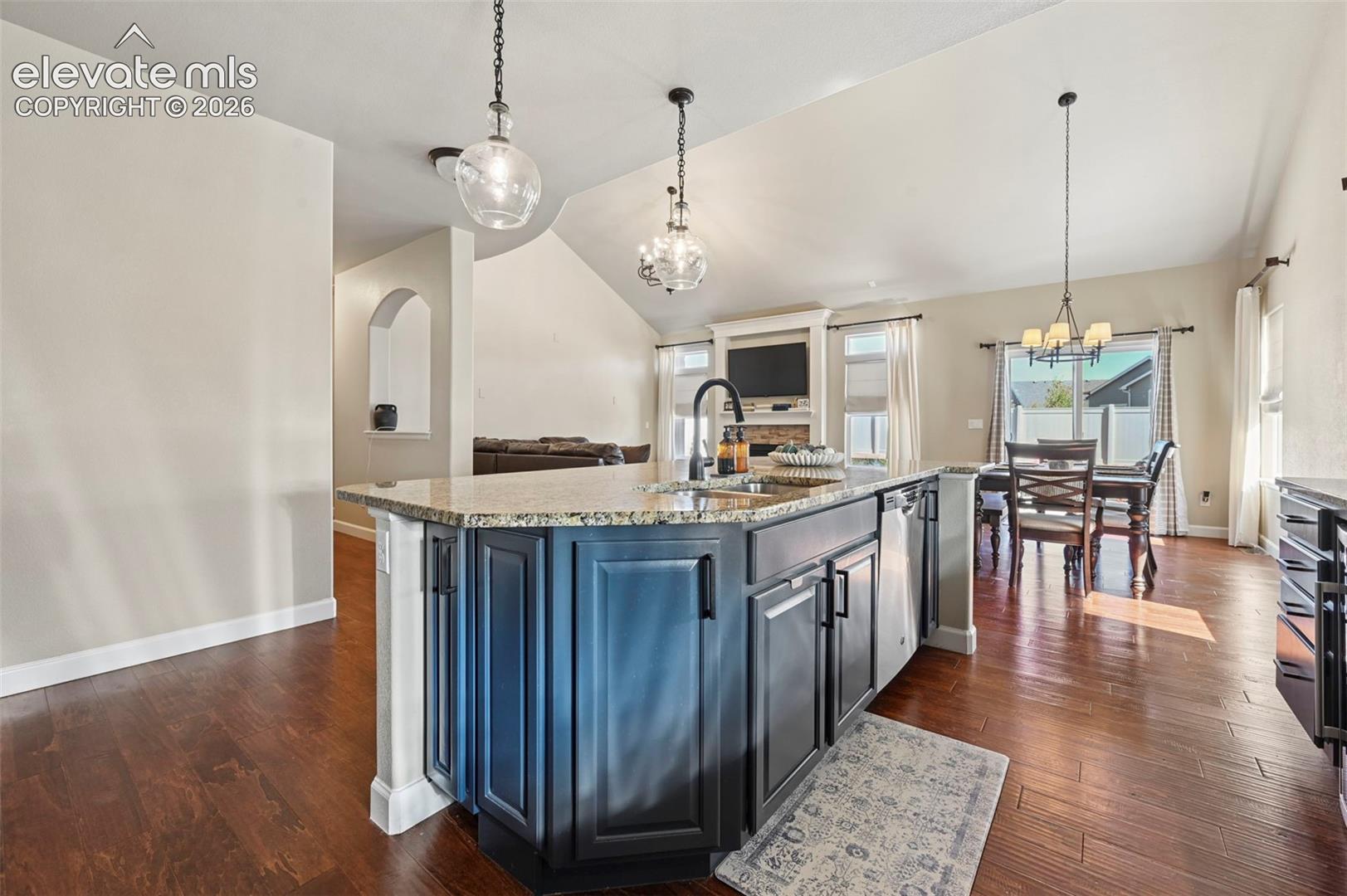 Kitchen island with dark cabinetry, stone countertops, and an undermount sink, complemented by wood-finish flooring, vaulted ceilings, and multi-light pendant fixtures