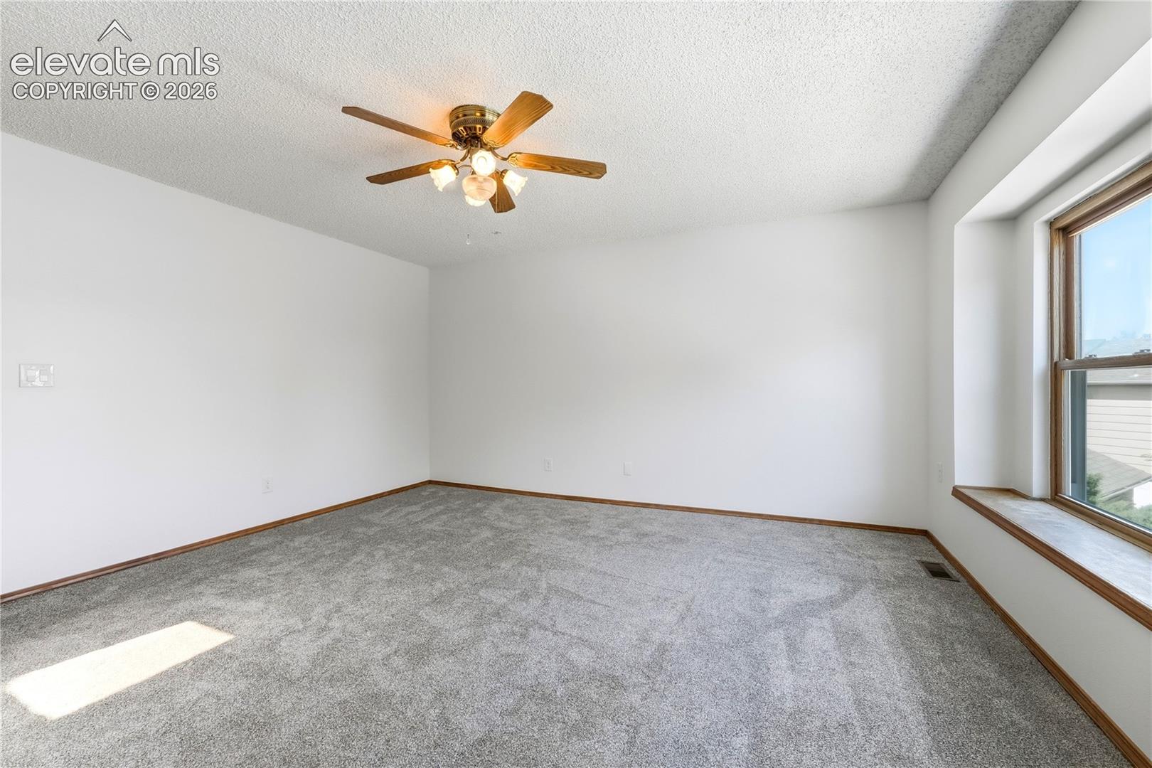 Spacious room featuring light gray carpeting, a wide window with faux wood vinyl trim, and a ceiling fan with lighting