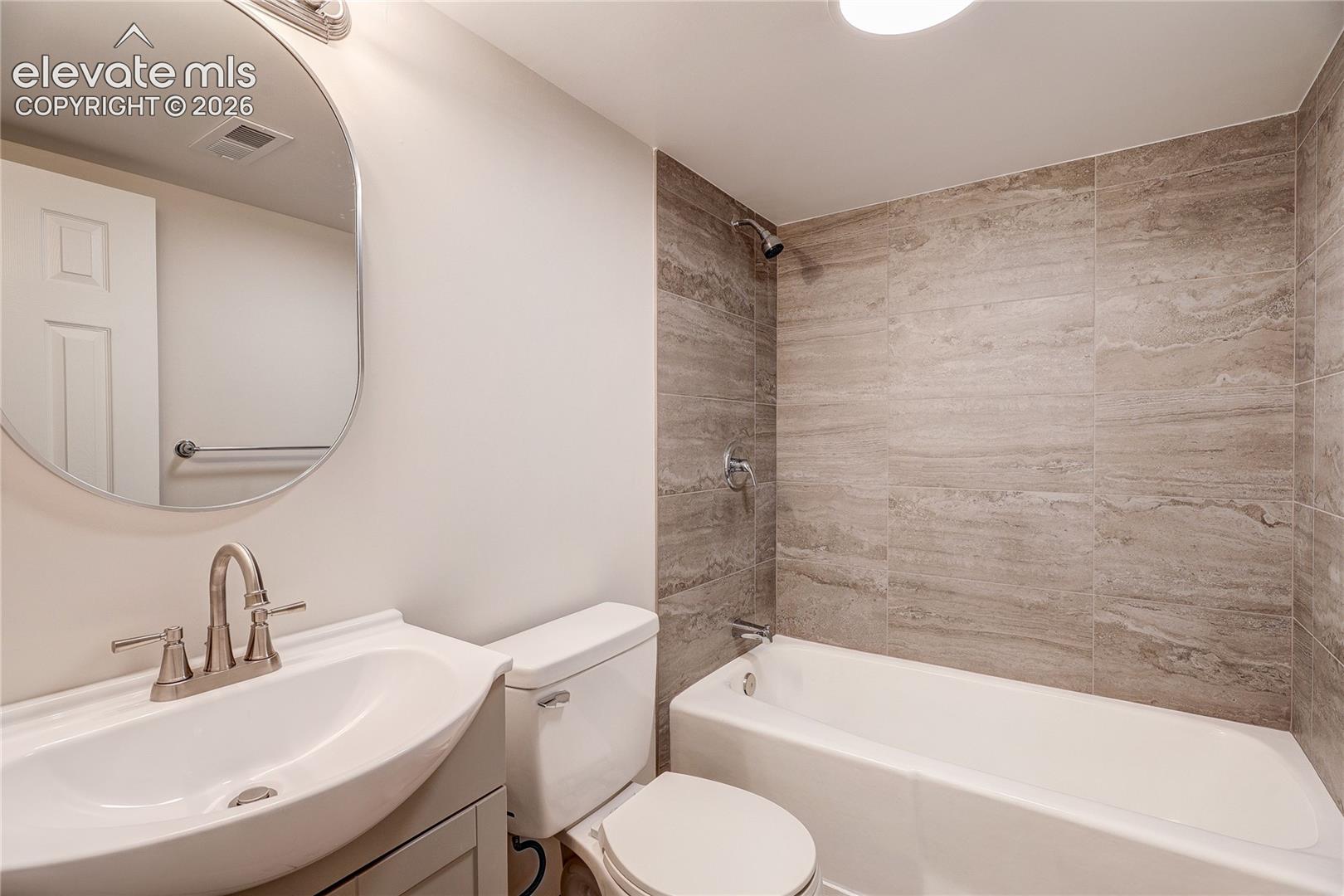 Bathroom featuring a vanity with a white basin sink, oval mirror, wall-mounted brushed metal hardware, and a tub enclosure with large-format textured stone-style wall tile