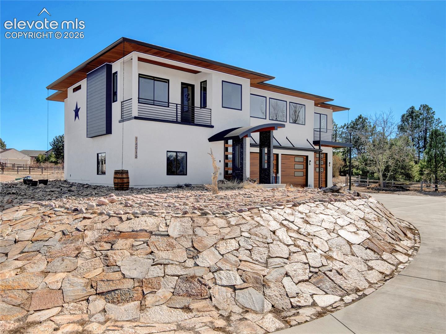Contemporary stucco exterior featuring a stone retaining wall, oversized windows, multiple balconies with dark railings, and wood-finish garage doors