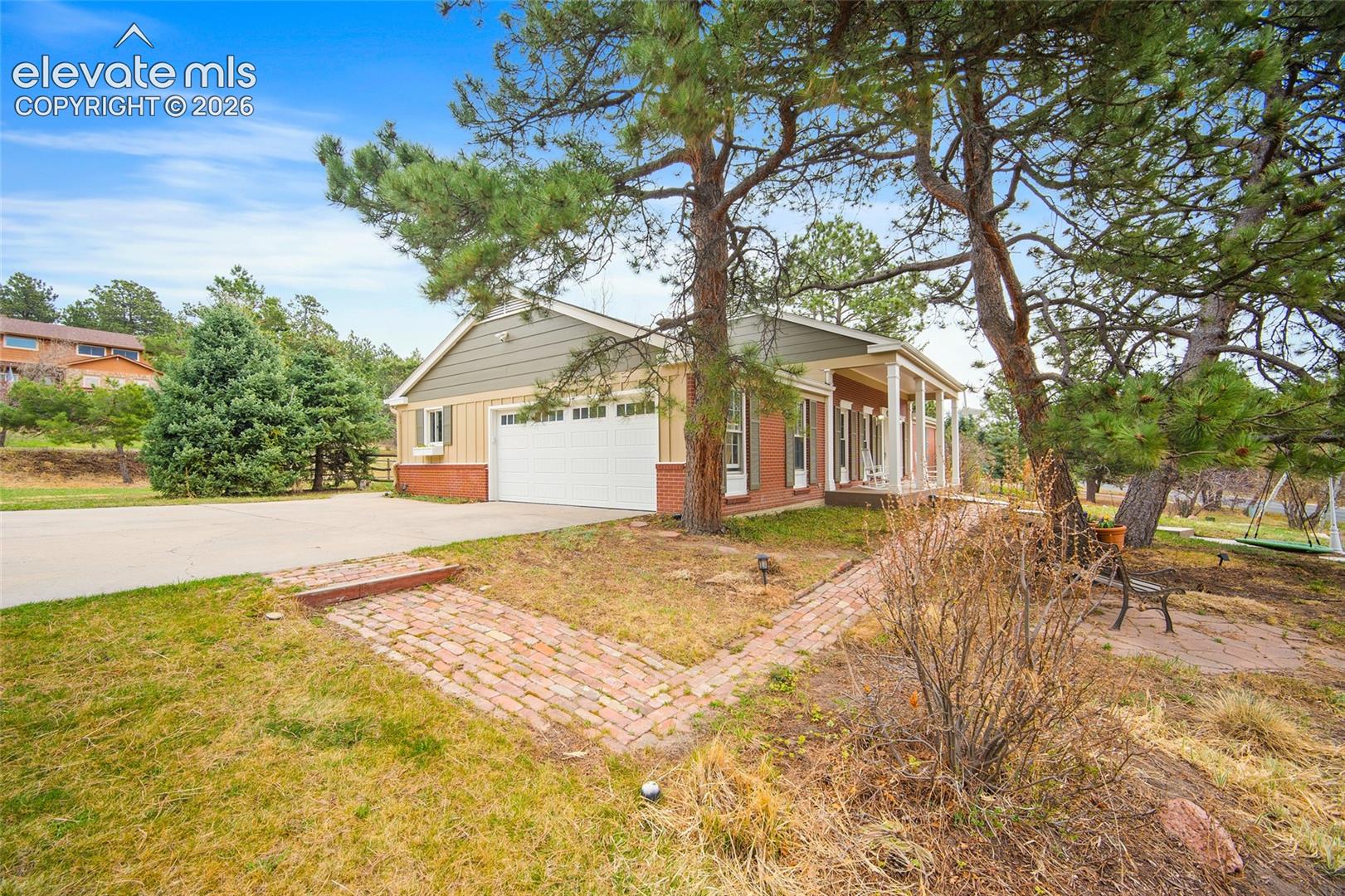 Single-story exterior featuring a brick and siding facade, an attached two-car garage, a covered front porch, and brick paver walkways surrounded by mature pine trees