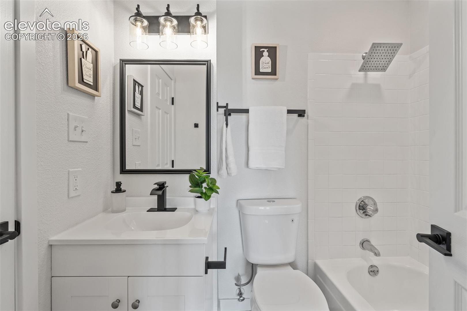 Bathroom featuring a vanity with a white countertop and matte black faucet, rectangular mirror with a black frame, wall-mounted three-light fixture with glass shades, and a tub-shower combination with white wall tile and a rainfall showerhead