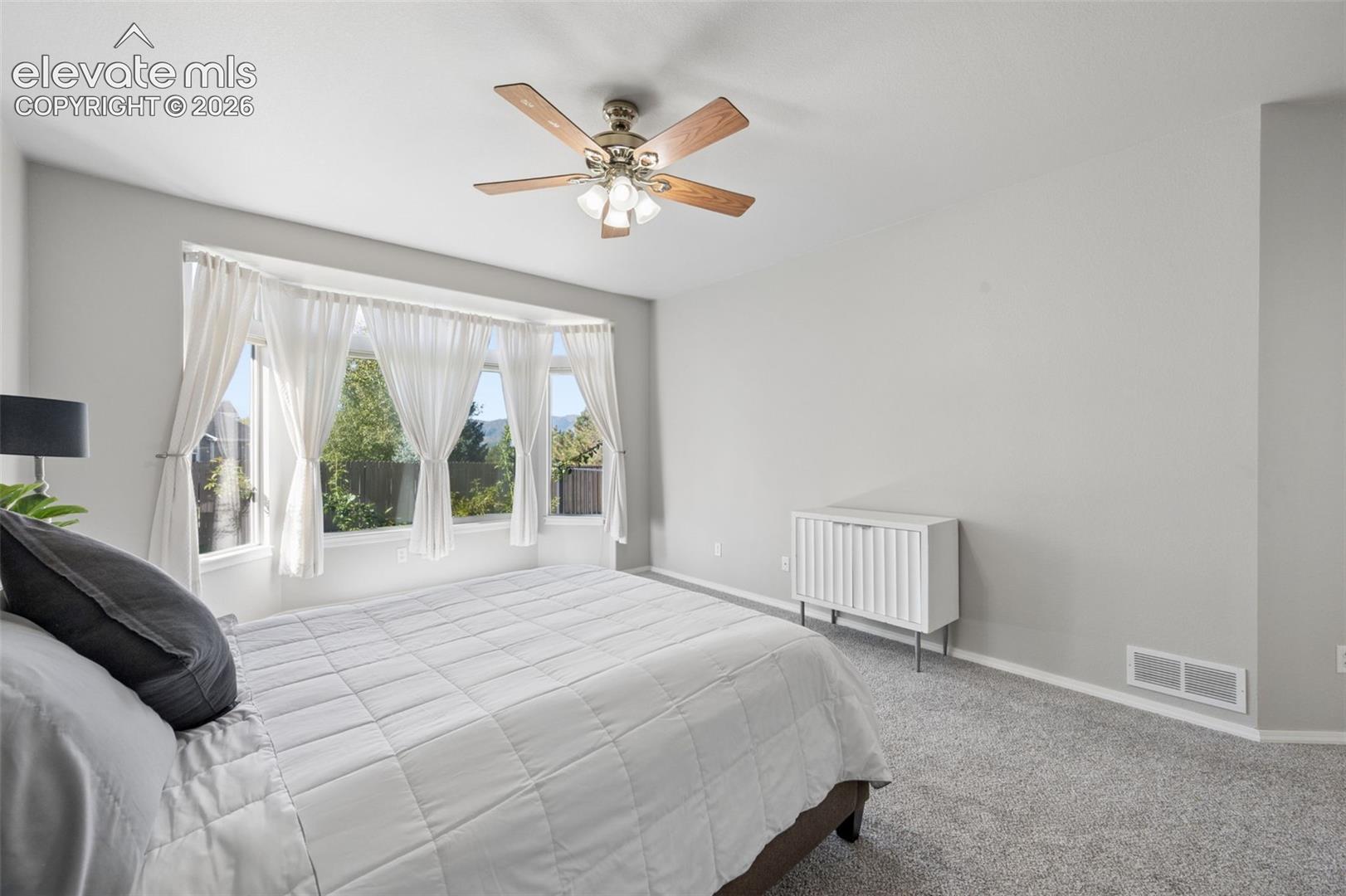 Bay window providing natural light, neutral interior wall paint, and a ceiling-mounted light fixture with a multi-blade fan