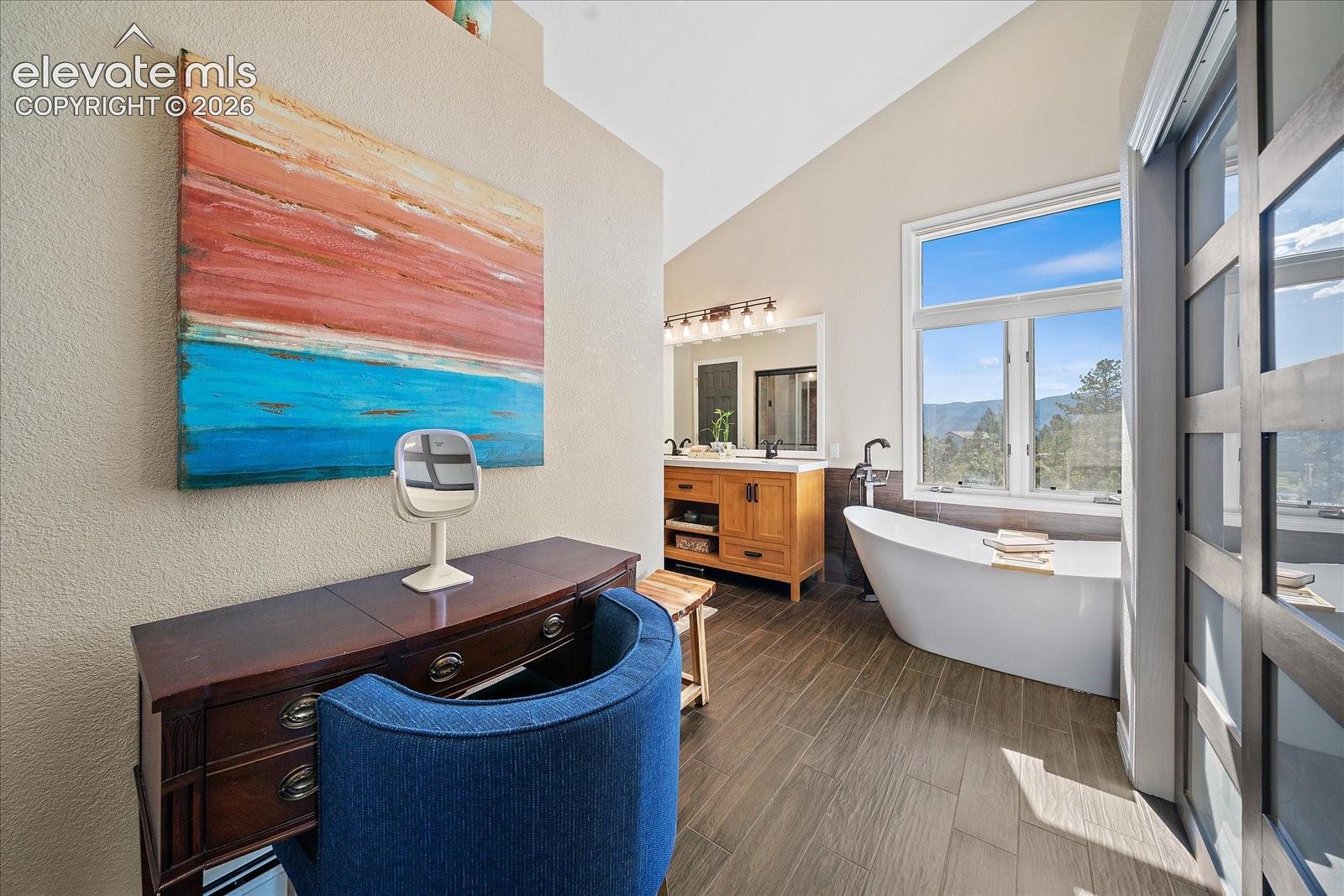 Vaulted ceiling, freestanding soaking tub, wood-finish flooring, wood cabinetry with dual sinks, and large picture window