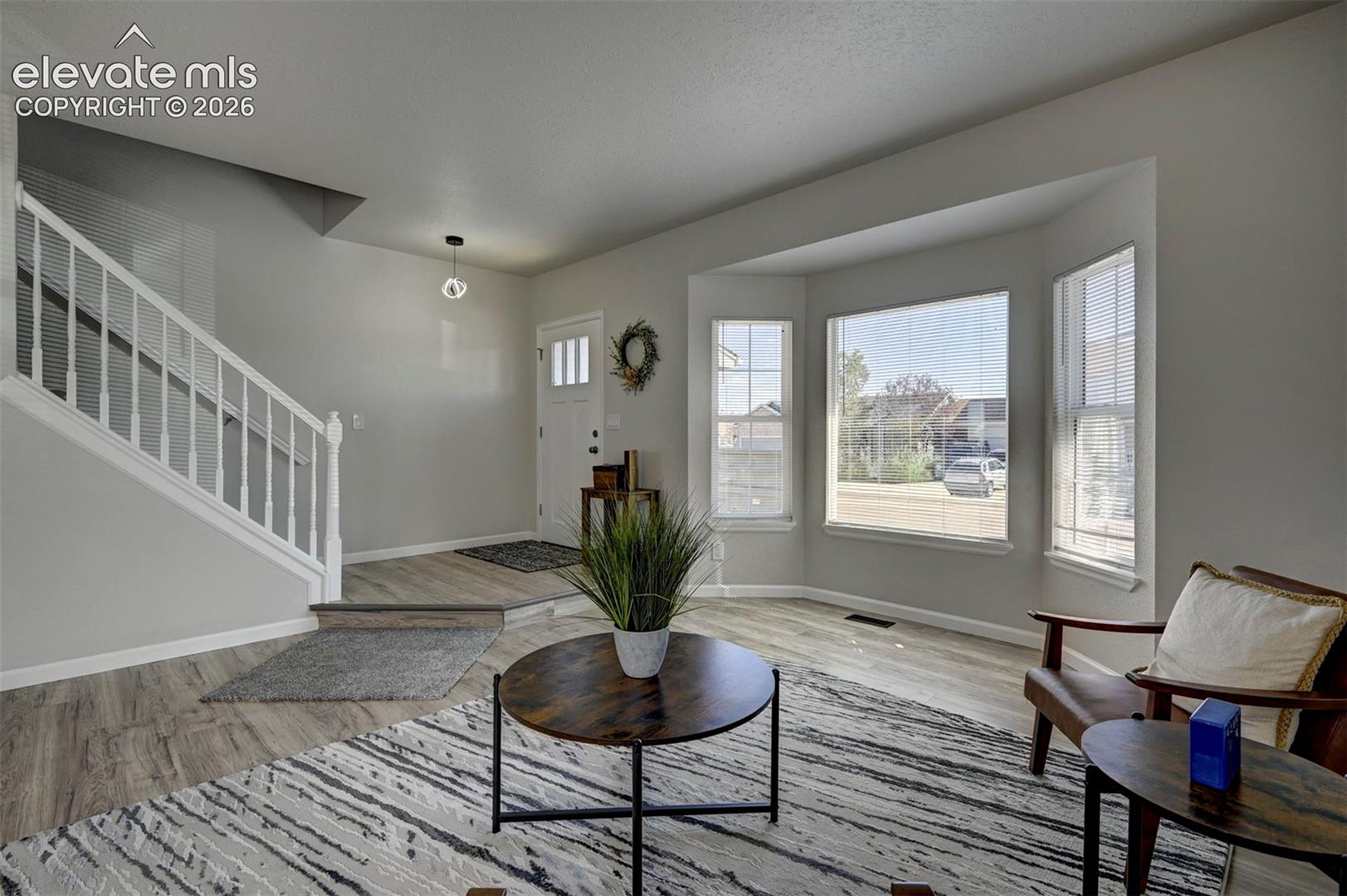 Foyer and living area featuring wood-finish flooring, a white staircase with a wooden handrail, a multi-panel entry door, and a large bay window