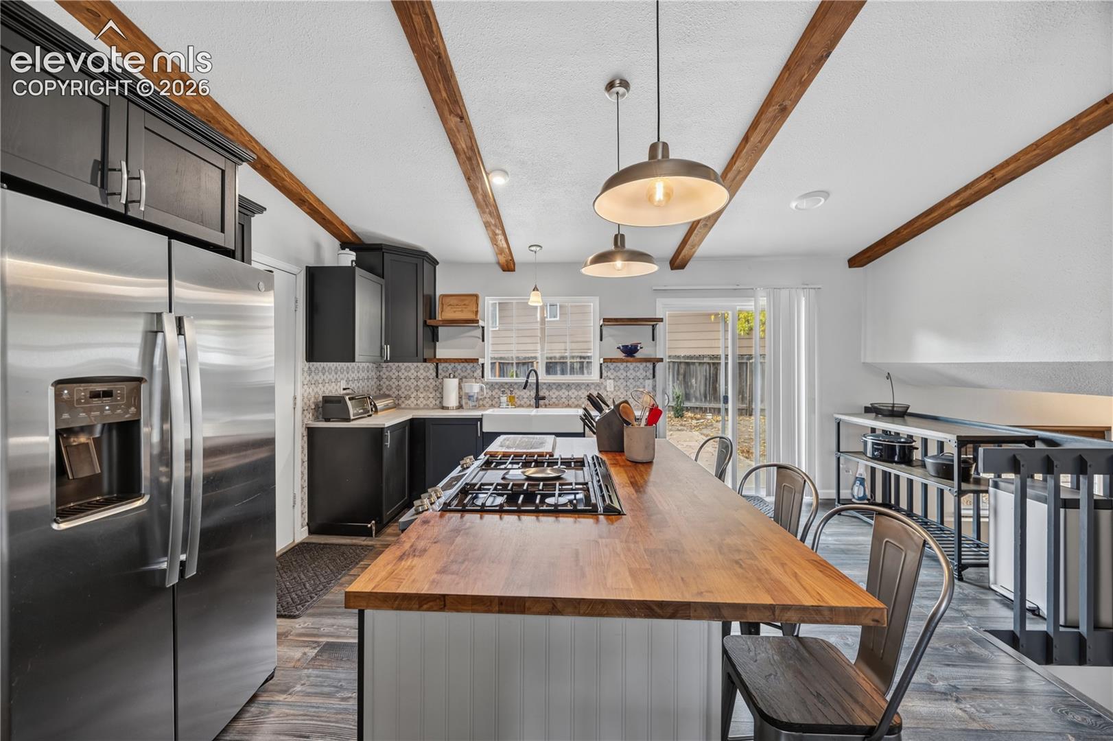 Kitchen featuring exposed ceiling beams, a wood-topped island with an integrated gas cooktop, farmhouse-style apron sink, custom cabinetry, and patterned tile backsplash