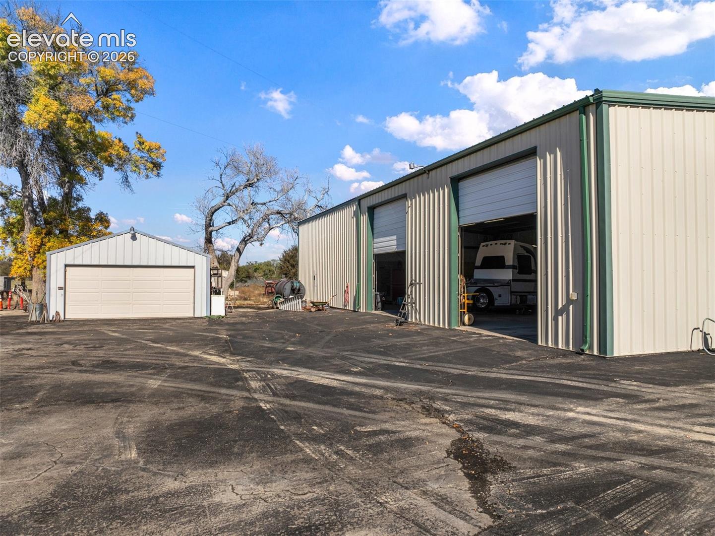 Large metal outbuilding featuring two expansive bays with roll-up doors, accompanied by a detached single-car garage and paved lot