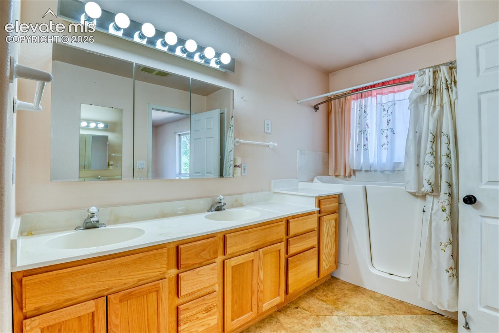 Dual-sink vanity with wood-finish cabinetry, solid surface countertop, large wall-mounted mirror, multi-bulb light fixture, and integrated walk-in tub