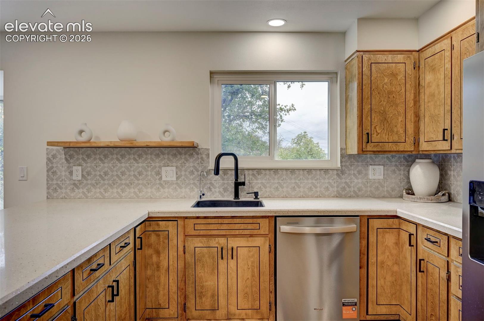 Kitchen featuring wood cabinetry with matte black hardware, stone-finish countertops, patterned tile backsplash, floating wooden shelving, and a matte black faucet