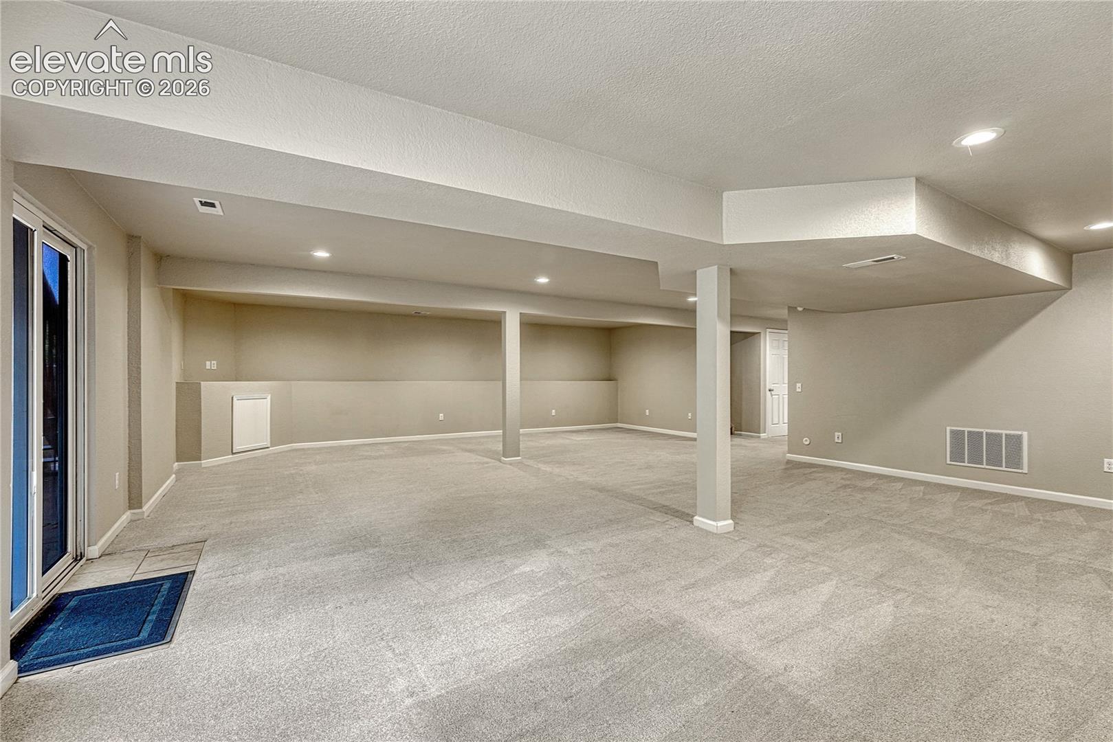 Open-concept finished space featuring neutral wall-to-wall carpeting, recessed ceiling lighting, a structural support column, and a sliding glass door with tile flooring inset