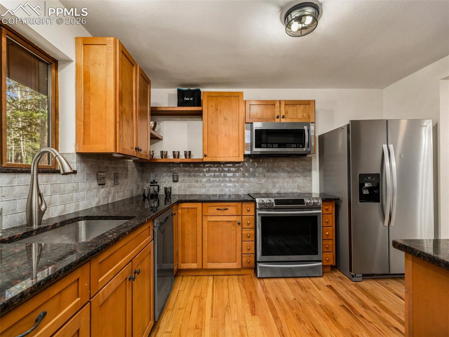 Kitchen with stainless steel appliances, open shelves, dark stone counters, light wood-style flooring, and wood finish cabinetry