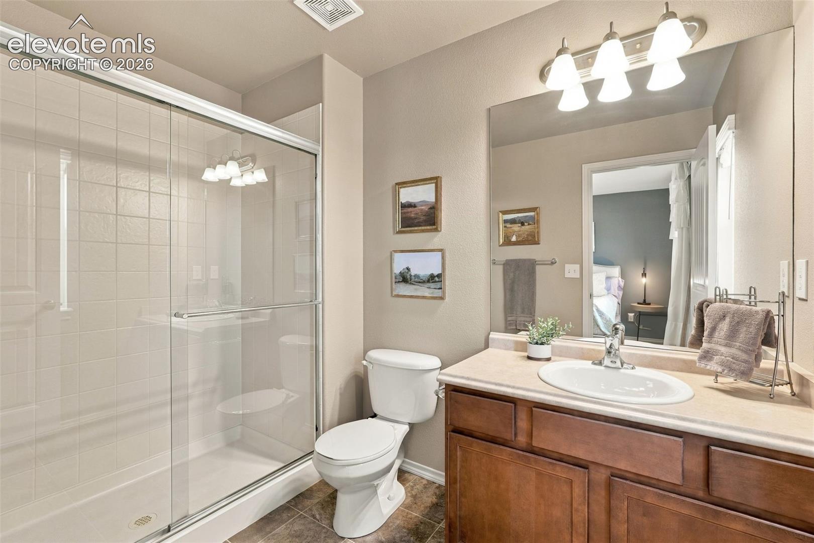 Bathroom featuring a wood-finish vanity with a stone-composite countertop, an integrated oval sink, a walk-in shower with a sliding glass enclosure, and a full-width vanity mirror with a three-light fixture
