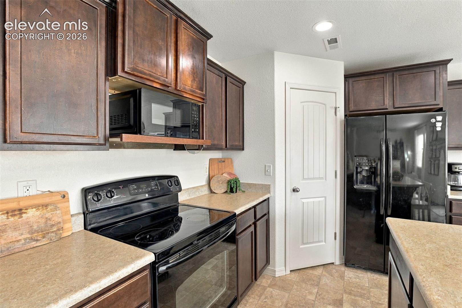 Kitchen featuring dark wood-finish cabinetry, light-toned countertops, black appliances, and a pantry door