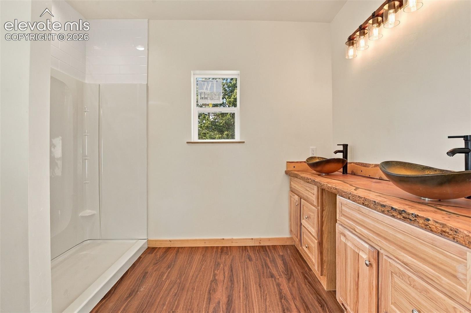 Bathroom featuring a dual-sink vanity with live-edge countertop and vessel sinks, matte black fixtures, wood-finish flooring, and a walk-in shower surround