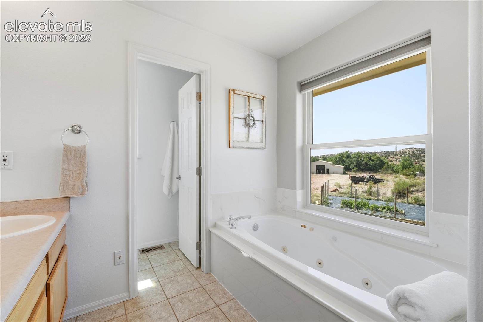 Soaking tub with integrated jets, large window with roller shade, light wood-finish vanity with countertop, and neutral tile flooring