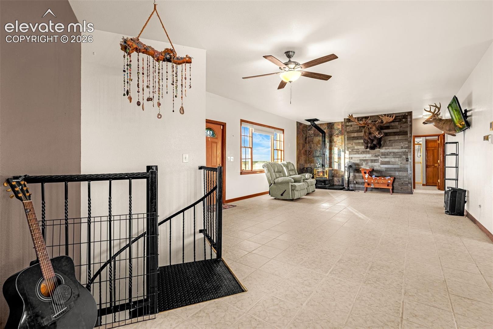 Spacious room featuring light-toned tile flooring, a wood-burning stove with a decorative wood-paneled wall, a ceiling fan with integrated lighting, and multiple windows