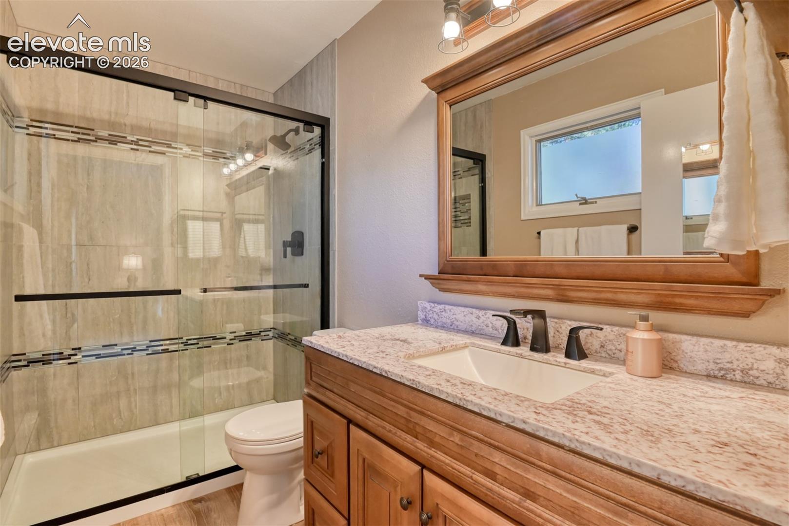 Primary bathroom vanity with a stone countertop with under-mount sink complemented by a large wood-framed mirror and a glass-enclosed shower with tiled surround