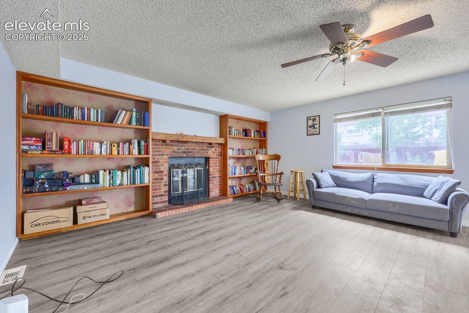 Light wood-finish flooring, built-in shelving, brick fireplace, and ceiling fan with integrated lighting
