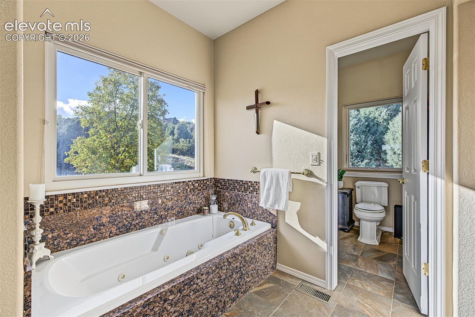 Primary bathroom featuring a jetted soaking tub with granite surround, tile flooring, and a large picture window offering mountain views.