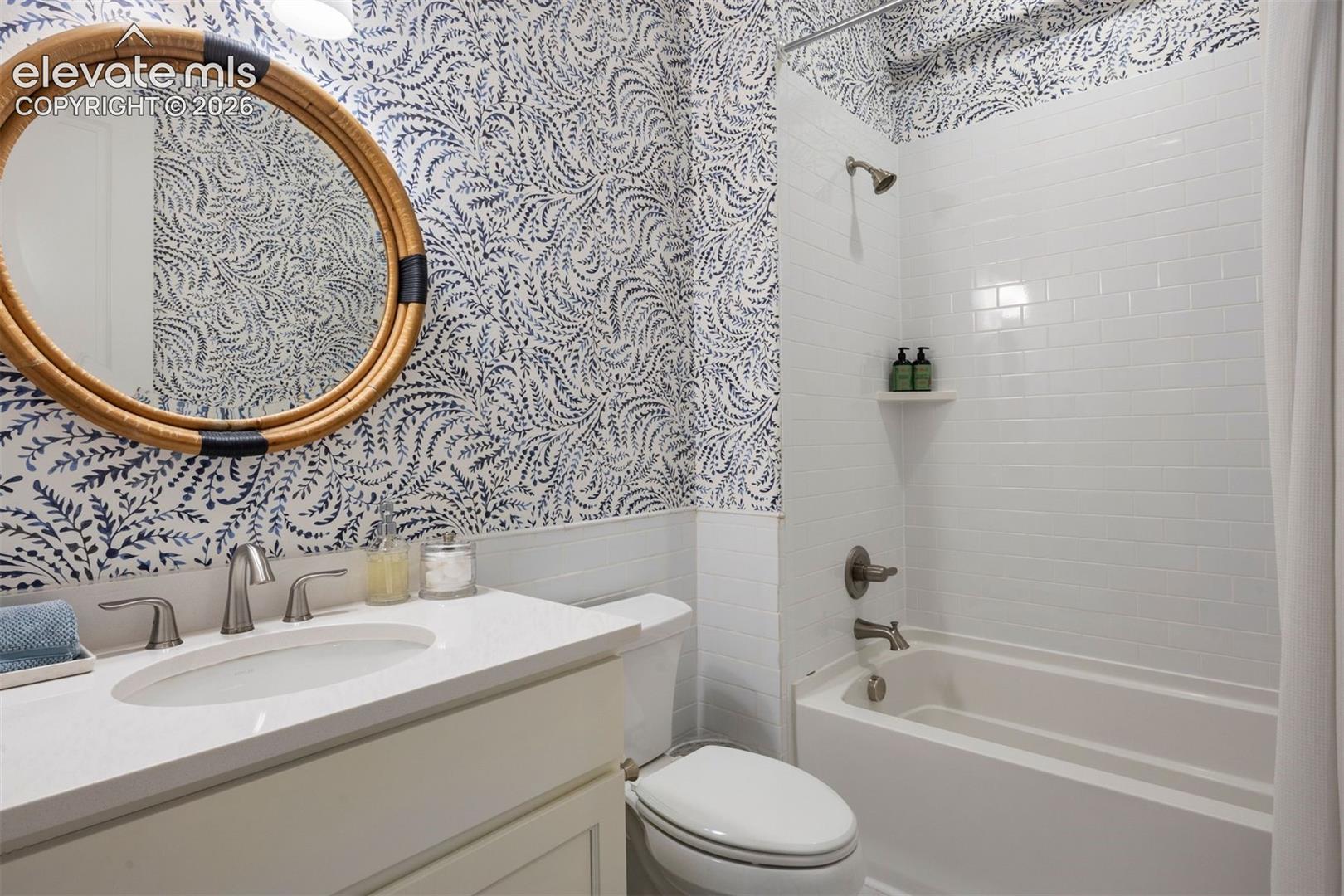 Bathroom featuring patterned wallpaper, a white vanity with an integrated sink, a round woven mirror, a white bathtub, and white subway tile surround