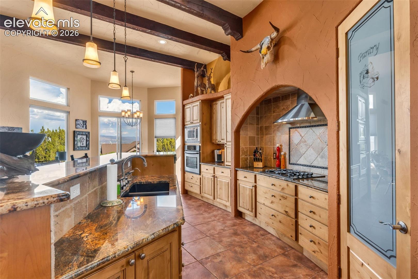 Kitchen featuring exposed timber ceiling beams, stone countertops, a decorative arched alcove over the cooktop, tile flooring, and a pantry door with frosted glass detailing