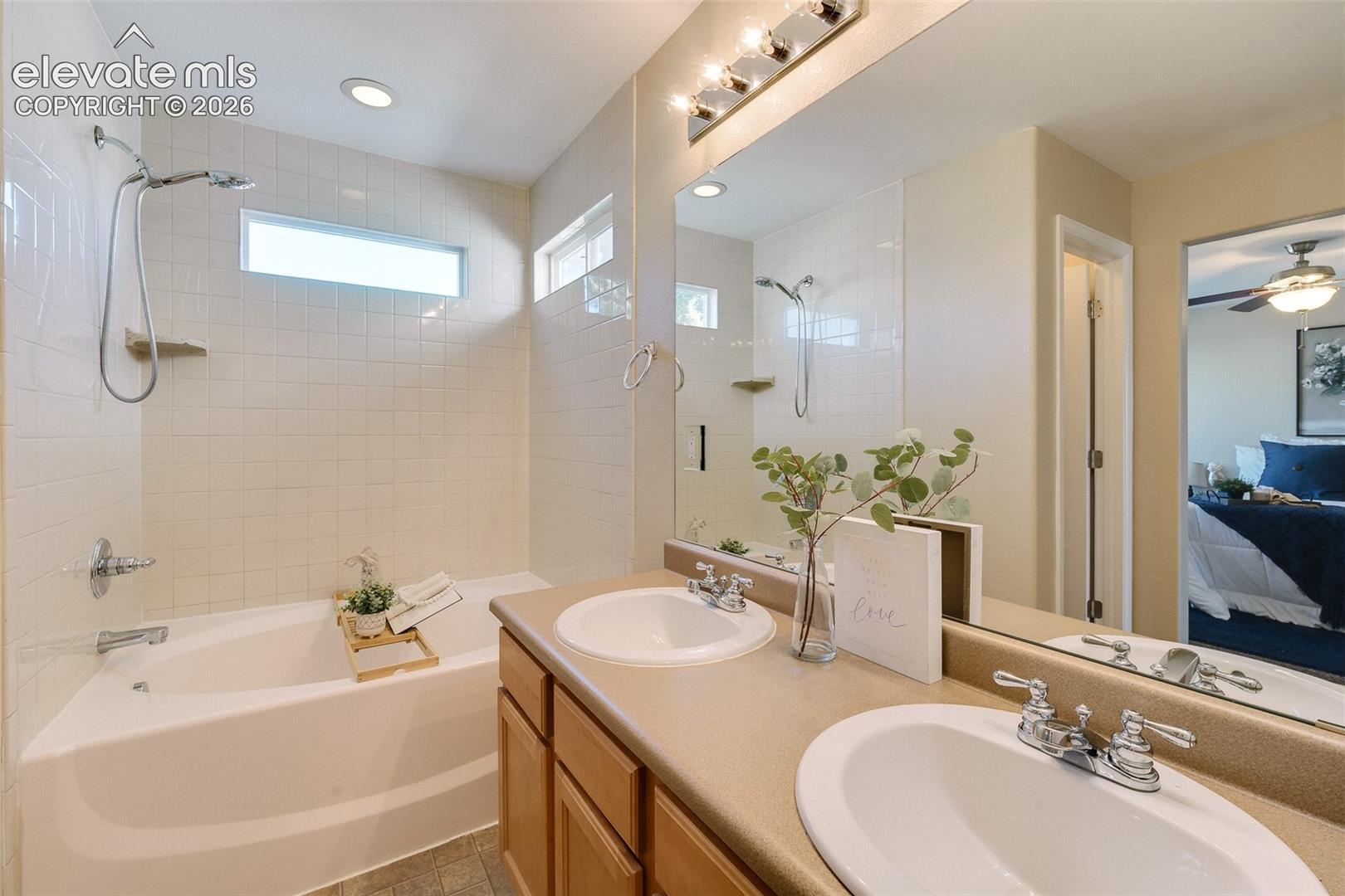 Dual-sink vanity with light-finish cabinetry, solid-surface countertop, chrome fixtures, integrated soaking tub with wall-mounted shower head, and full-width wall mirror
