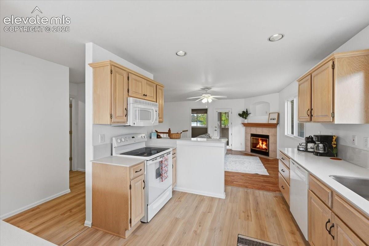 Kitchen featuring light-finish cabinetry, white appliances, and wood-finish flooring, incorporating a fireplace with a stone-finish surround