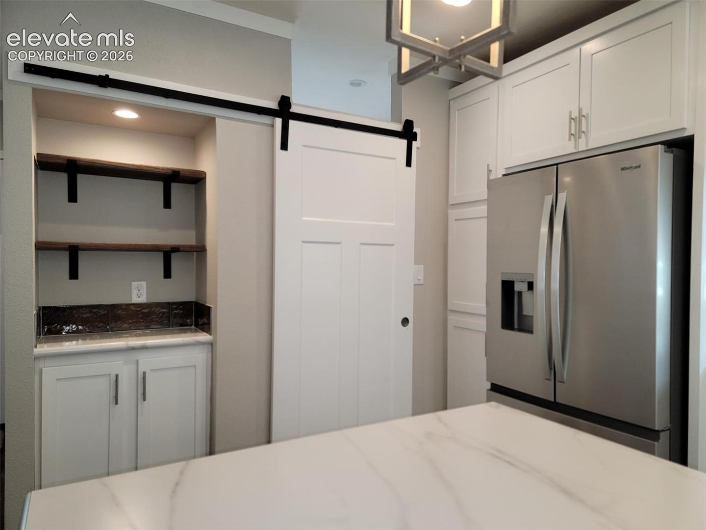 White shaker-style cabinetry, matte black sliding barn door hardware, stainless steel refrigerator, floating wood-finish shelving, and geometric overhead pendant lighting
