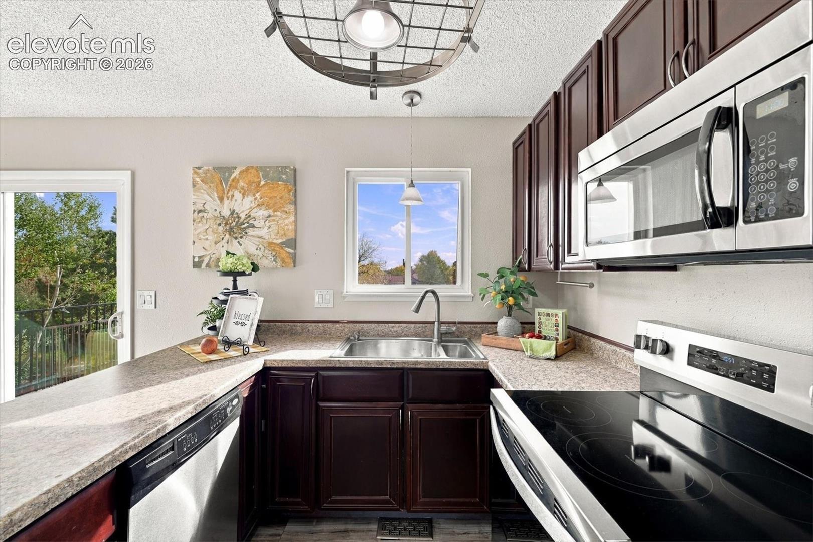 Kitchen featuring dark wood cabinetry, stainless steel appliances, a composite countertop, an undermount sink with a gooseneck faucet, and decorative metal pendant lighting