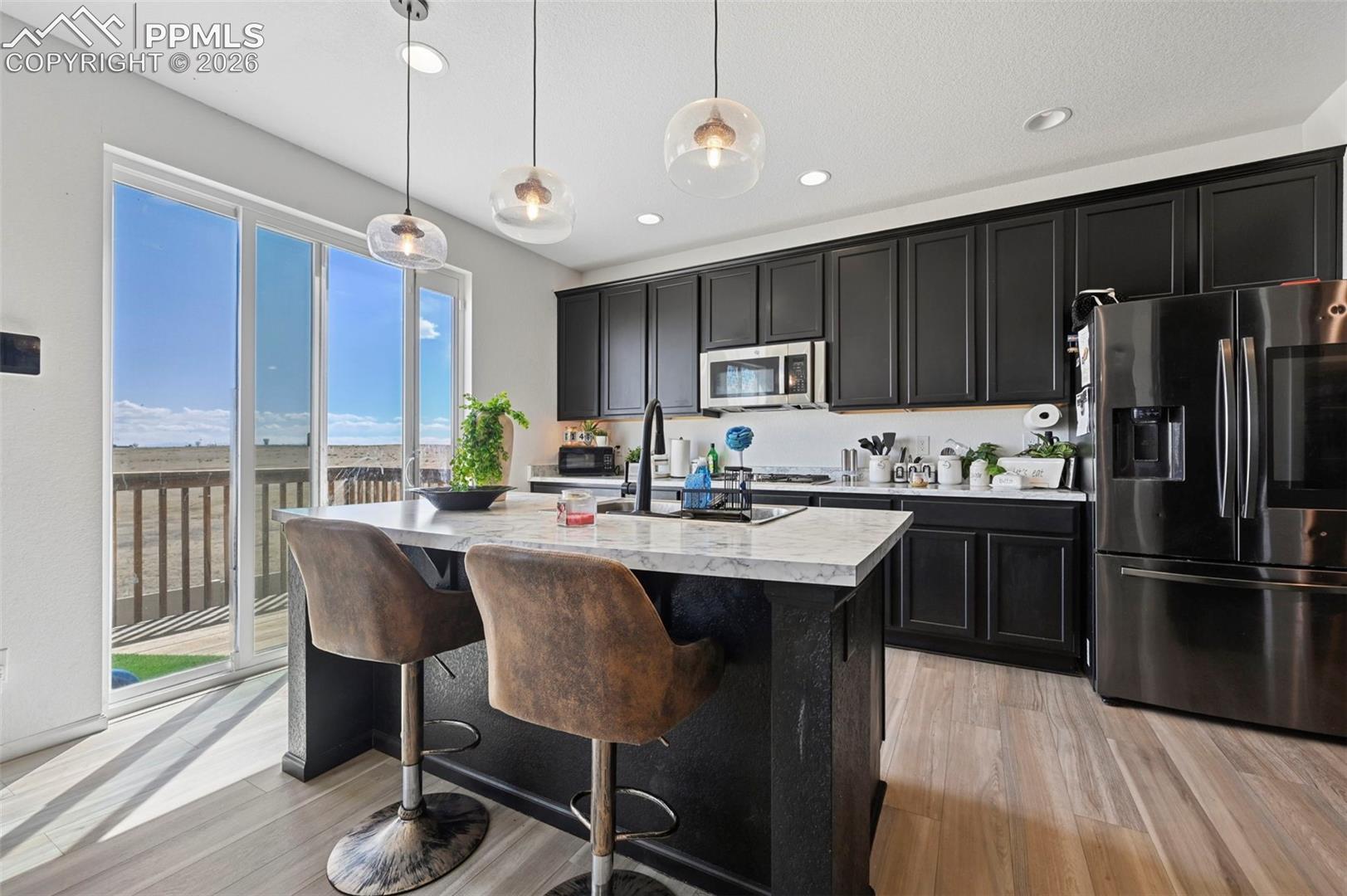 Contemporary kitchen featuring dark cabinetry, stainless steel appliances, a center island with pendant lighting, light-toned wood-finish flooring, and glass doors opening to an exterior deck