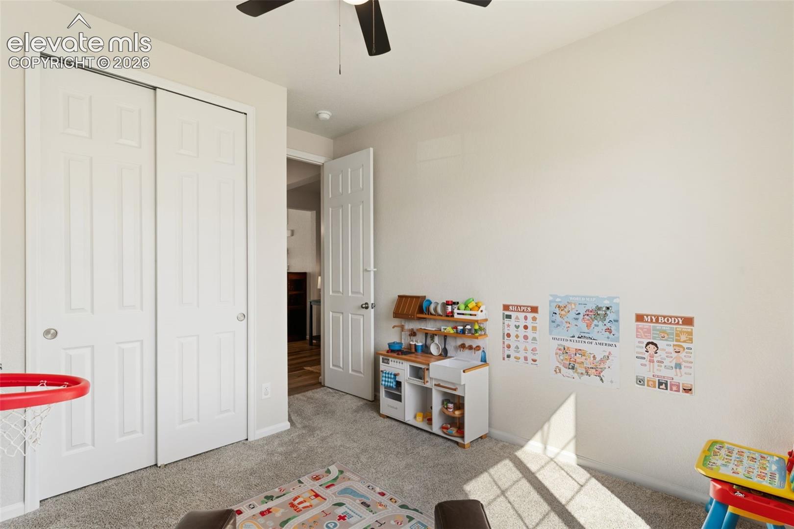 Neutral-toned interior room featuring light gray carpet, white walls, and a ceiling fan with dark blades