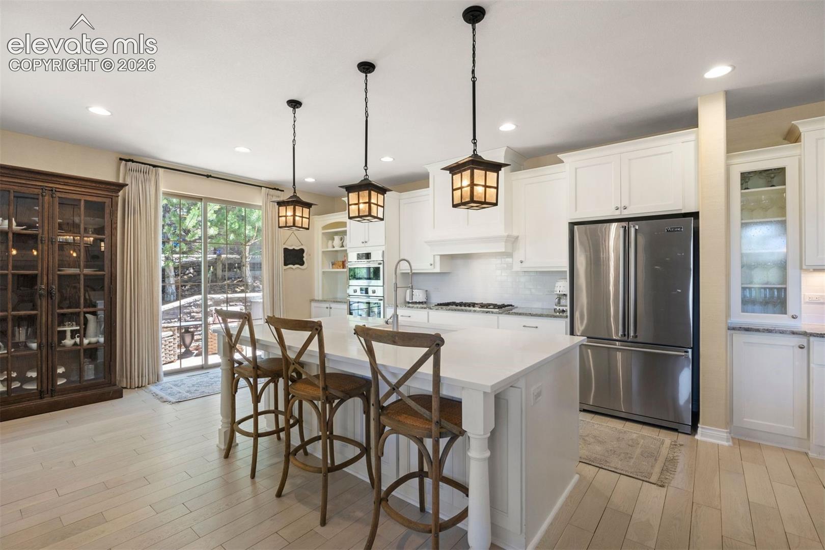Open-concept kitchen featuring an expansive central island with counter seating, light wood-finish flooring, white shaker cabinetry, stainless steel appliances, and elegant pendant lighting