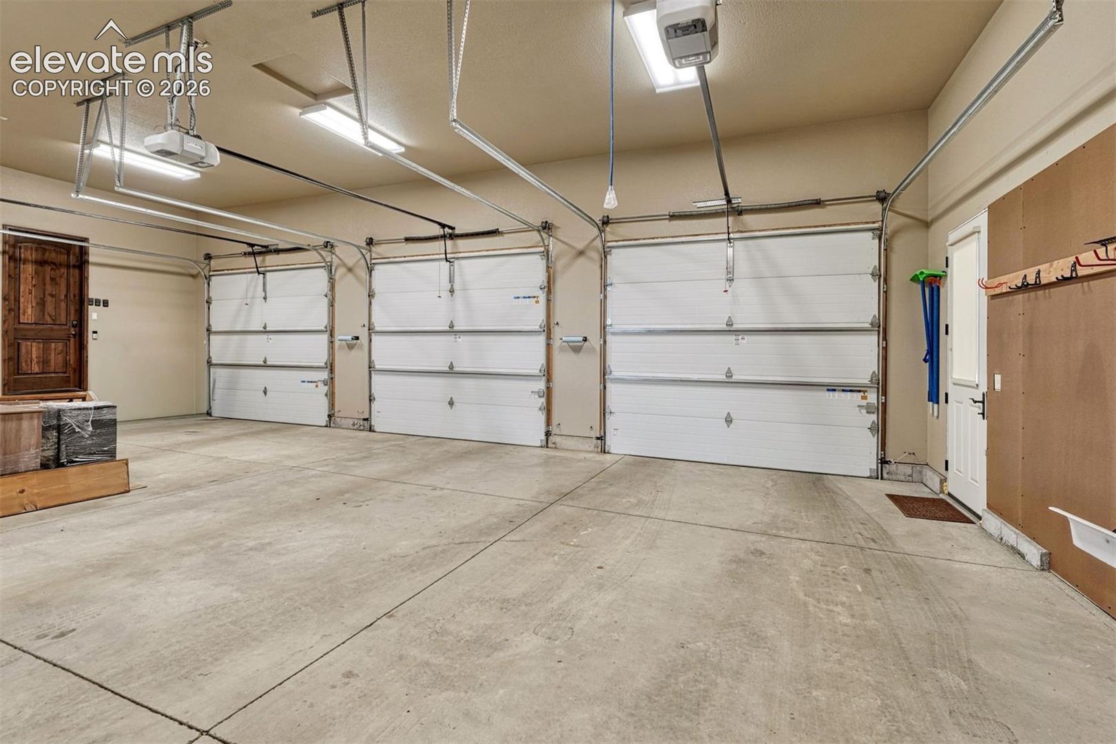 Expansive concrete floor garage with three overhead doors, overhead lighting, and a wood-finish entry door