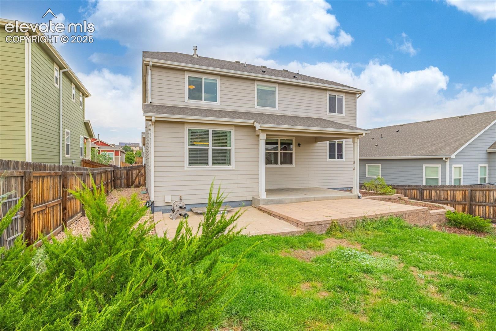Two-story exterior with light-toned siding, covered concrete patio, and an elevated paver patio with a low retaining wall