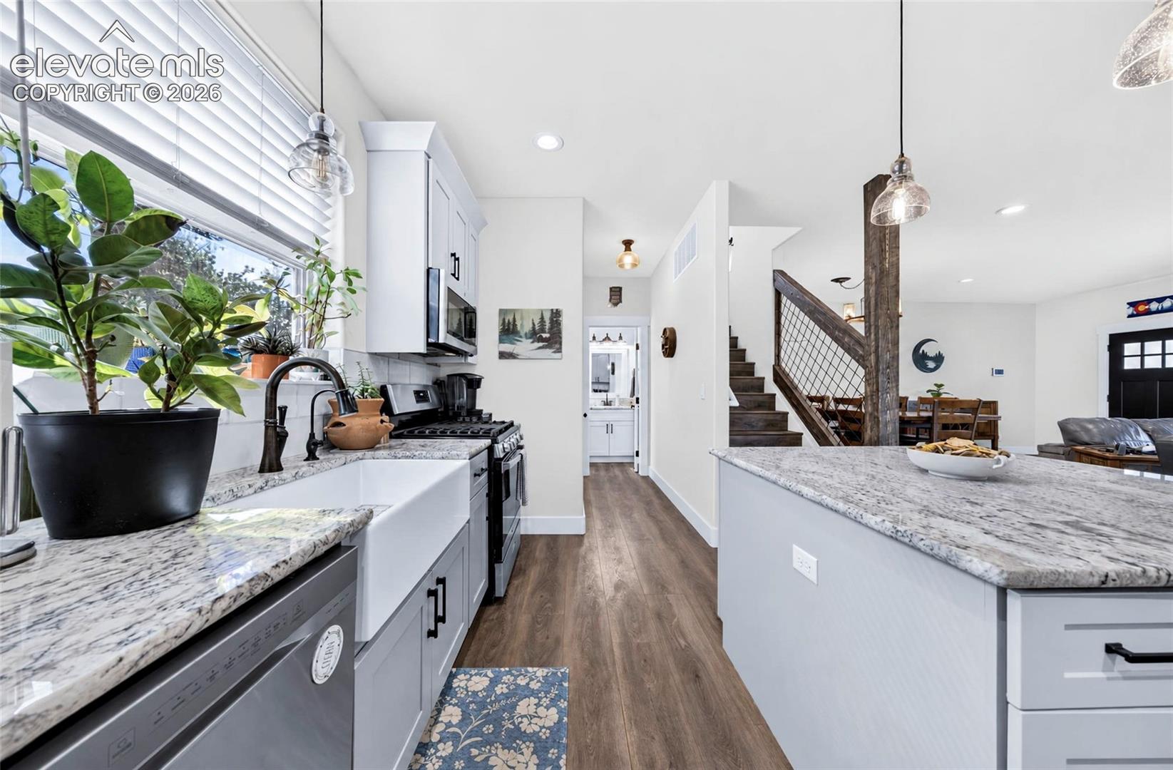 Kitchen featuring an expansive stone countertop island, apron-front farmhouse sink, wood-finish flooring, shaker-style cabinetry, and pendant lighting