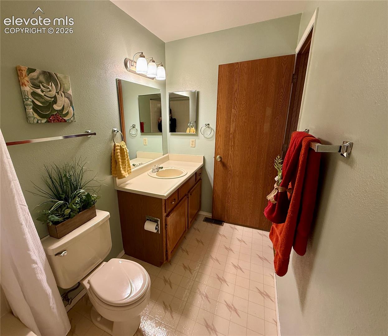 Bathroom featuring a wooden vanity with integrated countertop and sink, three-light wall fixture, large mirror, and vinyl flooring