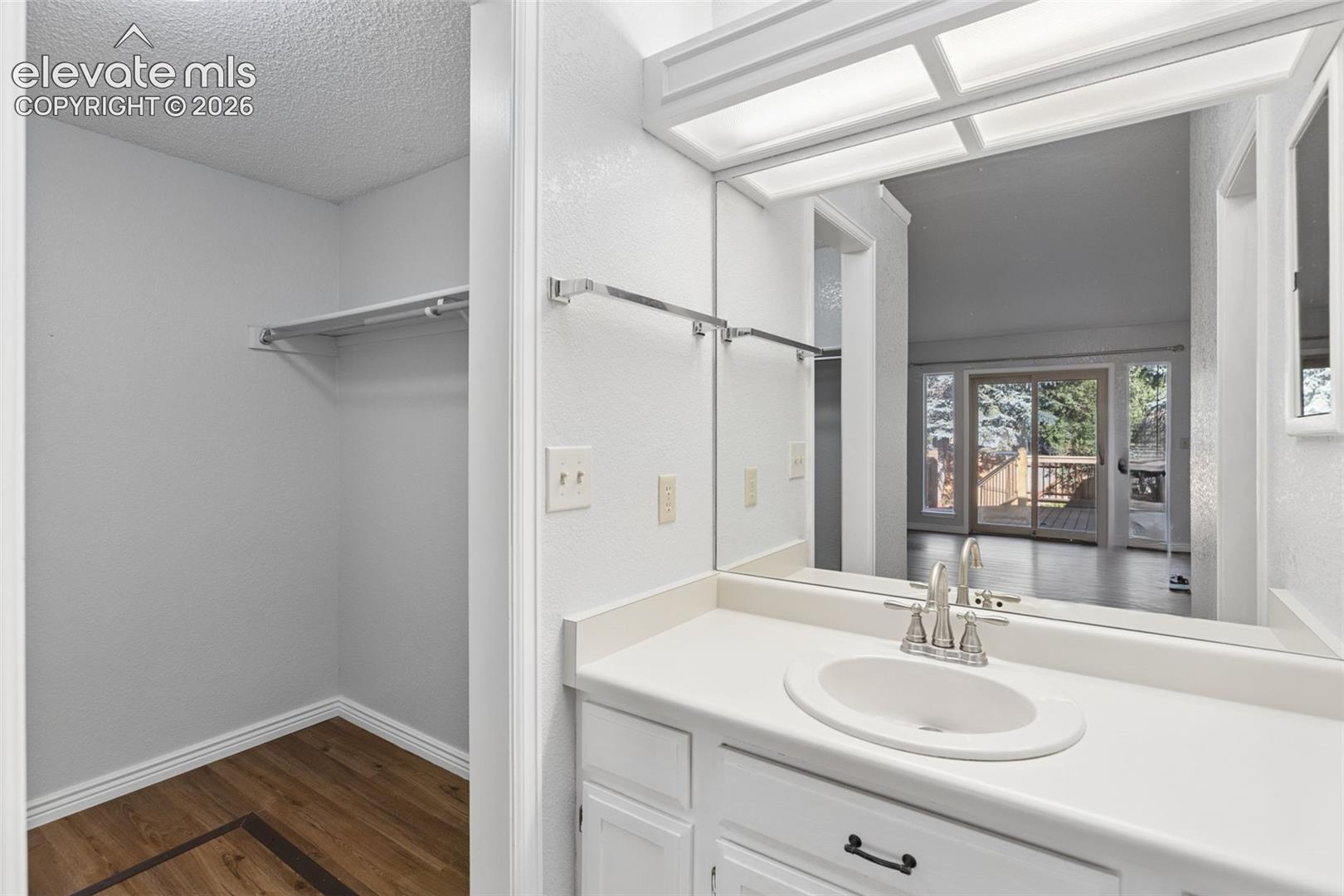 Vanity featuring a white countertop, oval undermount sink, chrome faucet, and white cabinetry with dark hardware, paired with a full-width wall mirror, overhead lighting, and an walk-in closet with wood-finish flooring