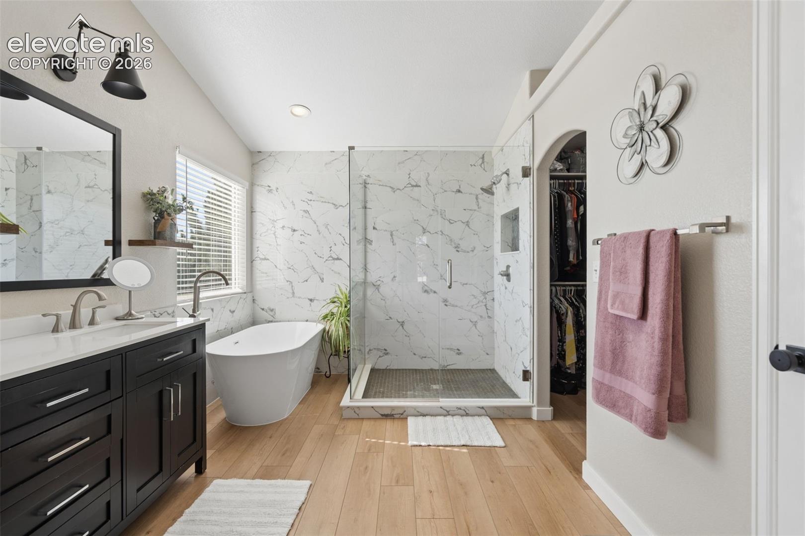 Spacious bathroom featuring a freestanding tub, glass-enclosed shower, luxury vinyl flooring, dark wood vanity with white countertop, and arched entry to a closet
