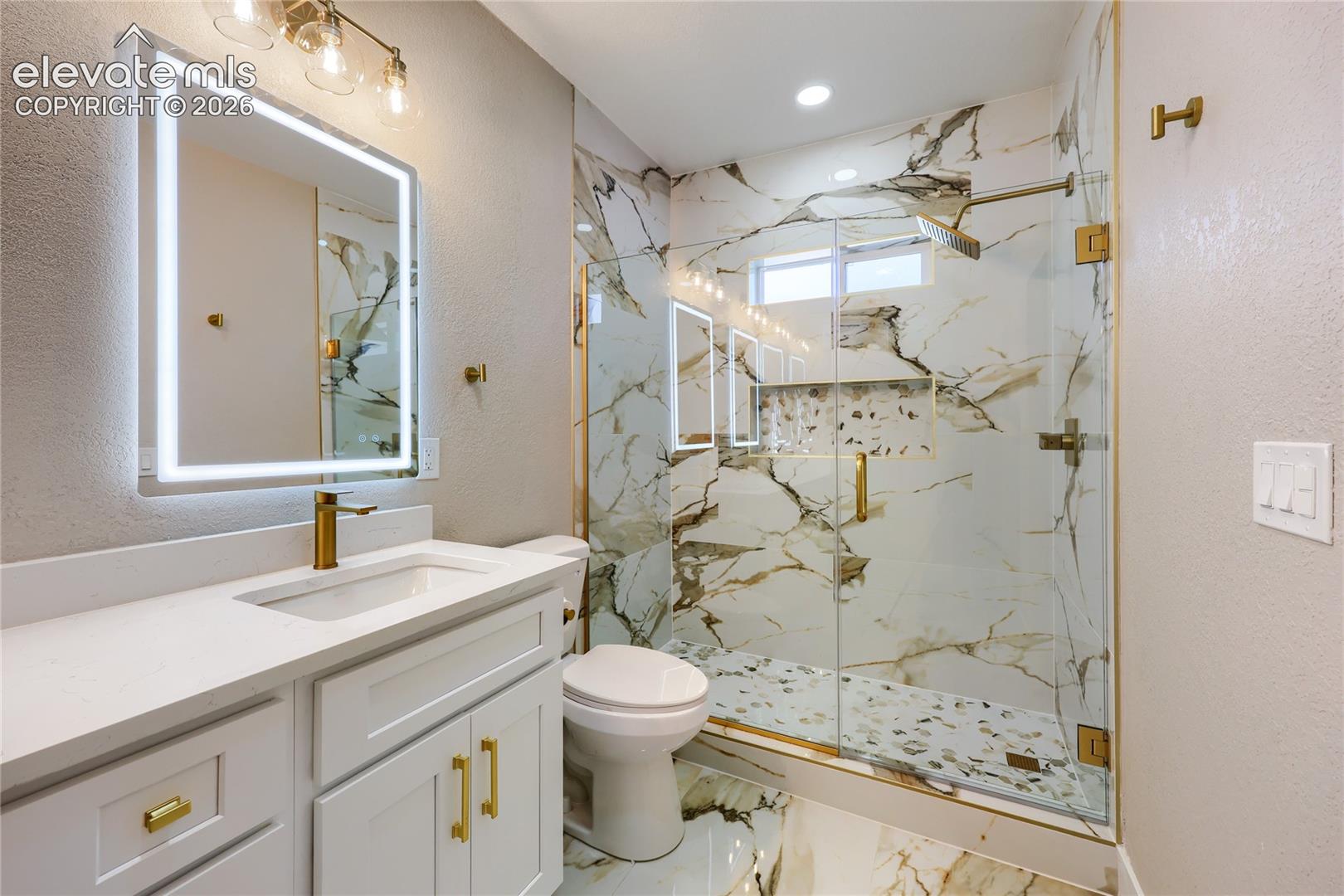 Spectacular 4-piece, spa-inspired primary ensuite bathroom with a marble shower surround and flooring, a quartz countertop, a custom-built shower niche and contemporary gold fixtures and finishes.