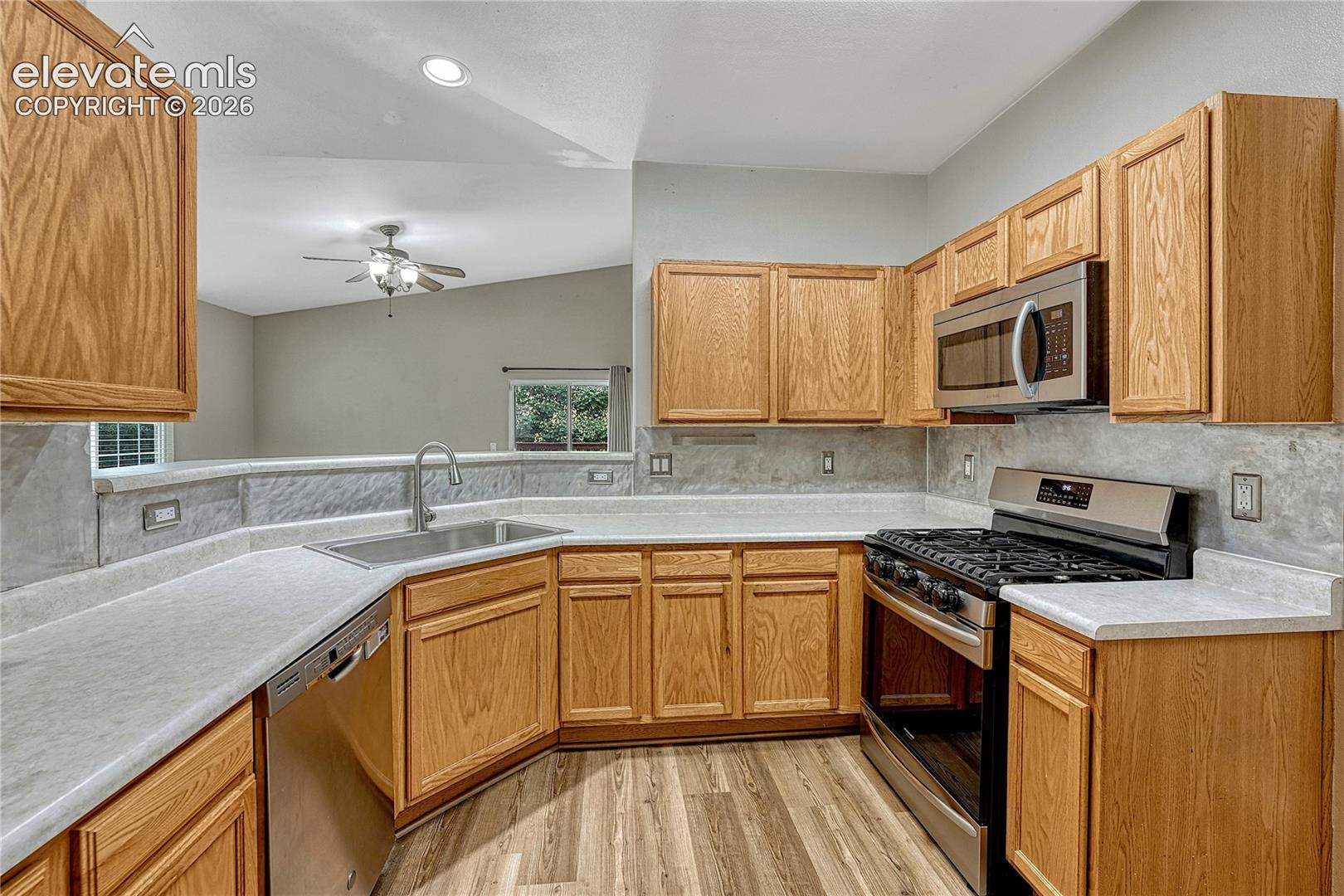 Kitchen featuring natural finish wood cabinetry, light-toned countertops, stainless steel appliances, a textured stone-finish backsplash, and wood-finish flooring