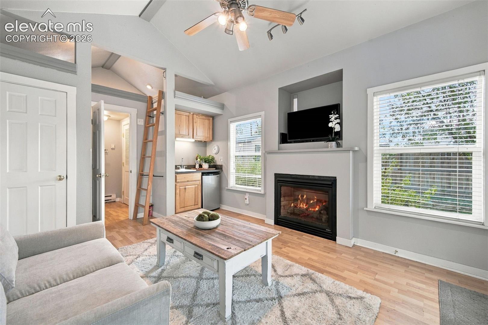 Vaulted ceiling, built-in fireplace with recessed media niche, wood-finish flooring, kitchenette with wood cabinetry, and integrated ceiling fan