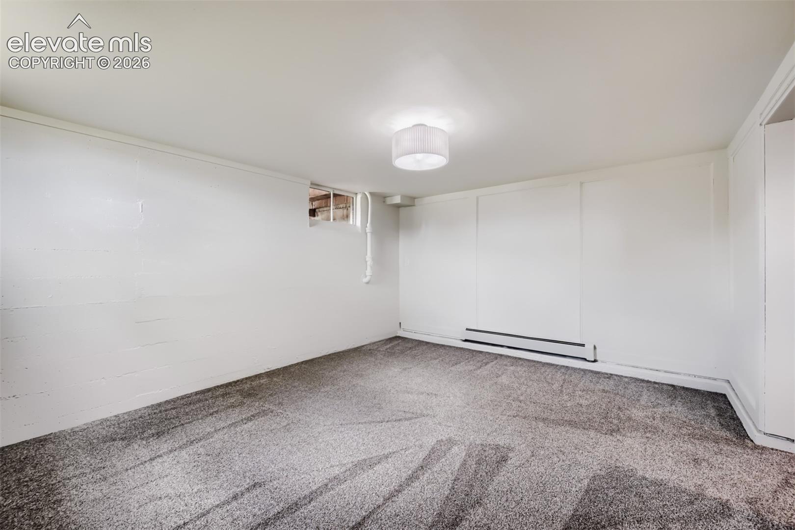 Textured gray carpeting, white painted walls, ceiling-mounted light fixture, and baseboard heating unit