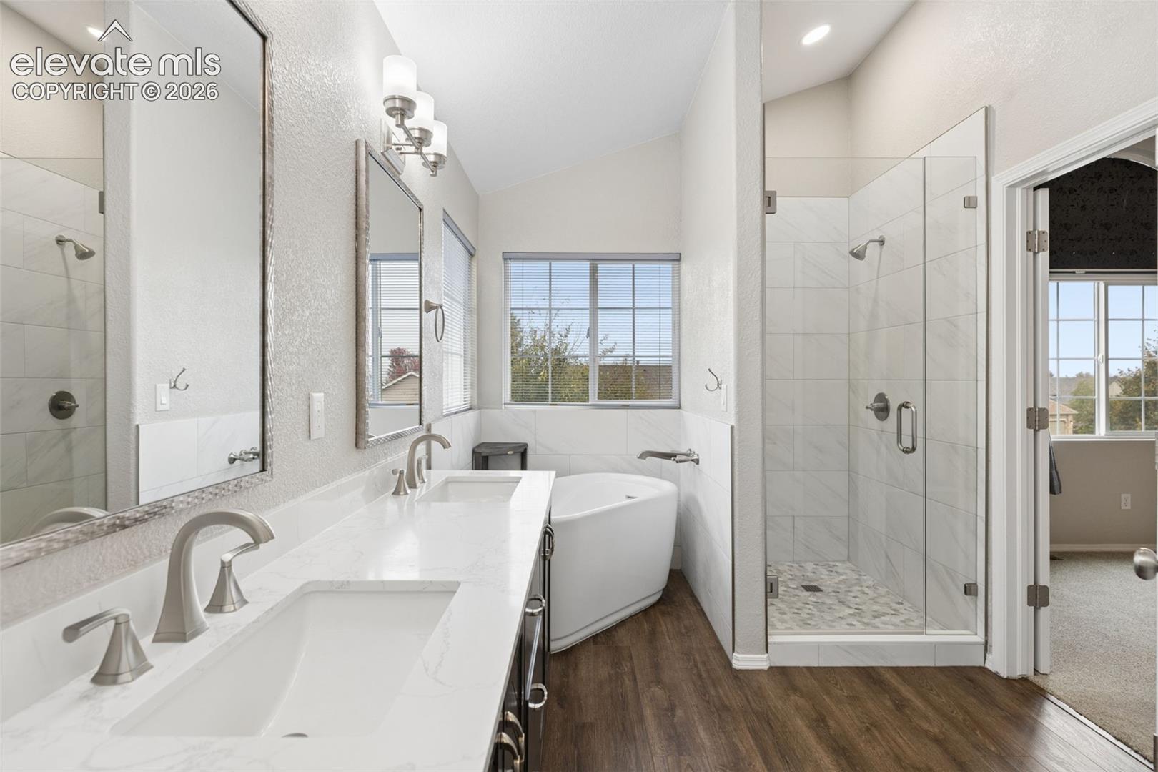 Bathroom featuring a double vanity with stone-style countertops, an enclosed glass-paneled shower with tiled surround, a corner soaking tub, and wood-finish flooring