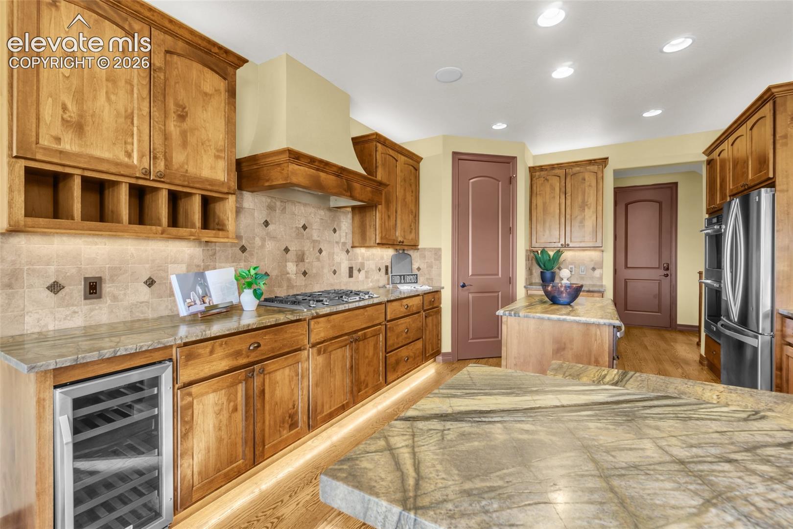 Kitchen featuring custom wood cabinetry, natural stone countertops, a tiled backsplash with decorative accents, built-in stainless steel appliances, and under-cabinet lighting